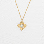 The Holly Yashi Petite Fleur Gold Necklace showcases a four-petal flower pendant with a sparkling Austrian crystal at the center, elegantly set against a white textured surface.
