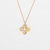The Holly Yashi Petite Fleur Gold Necklace showcases a four-petal flower pendant with a sparkling Austrian crystal at the center, elegantly set against a white textured surface.