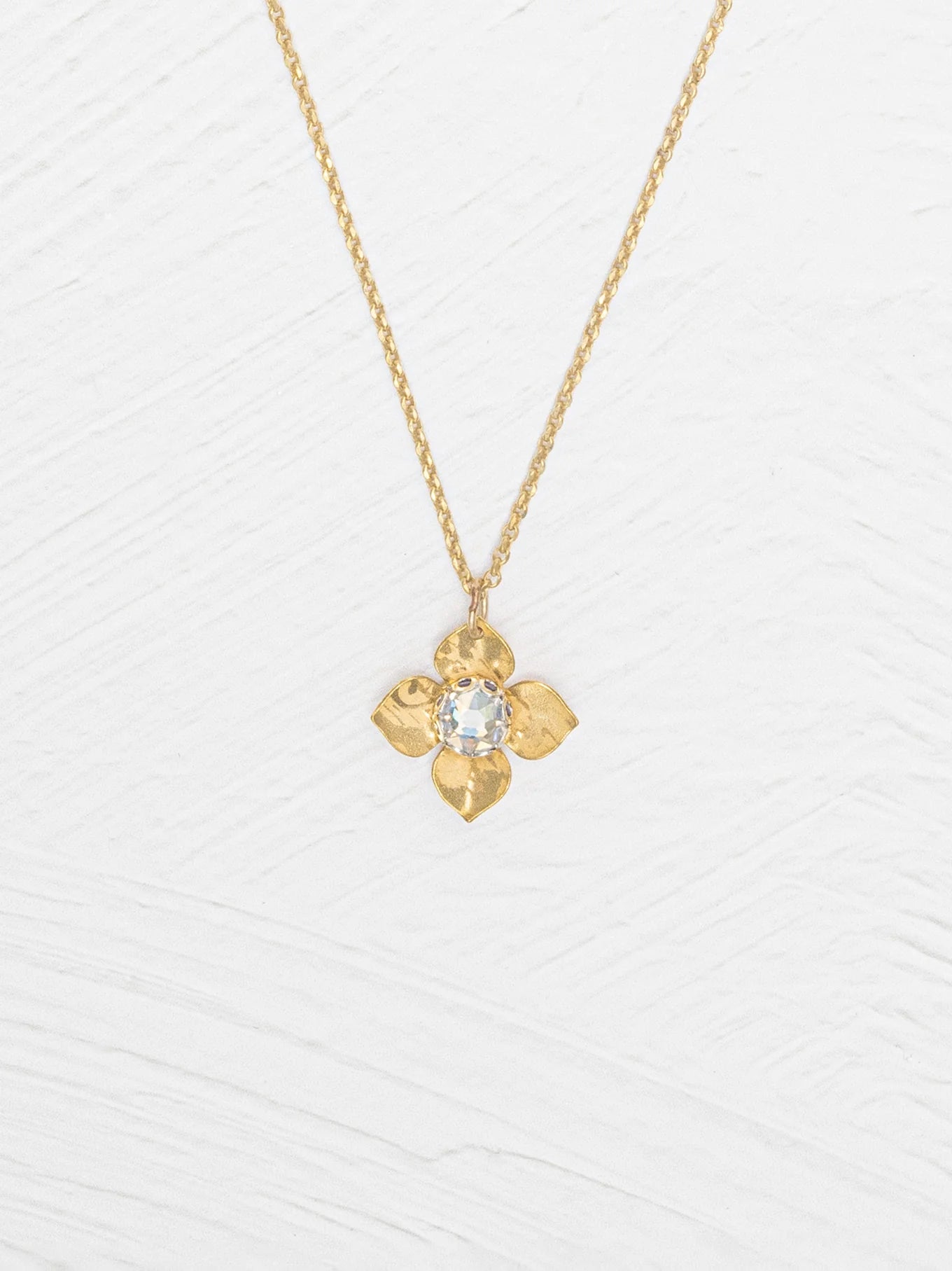 The Holly Yashi Petite Fleur Gold Necklace showcases a four-petal flower pendant with a sparkling Austrian crystal at the center, elegantly set against a white textured surface.