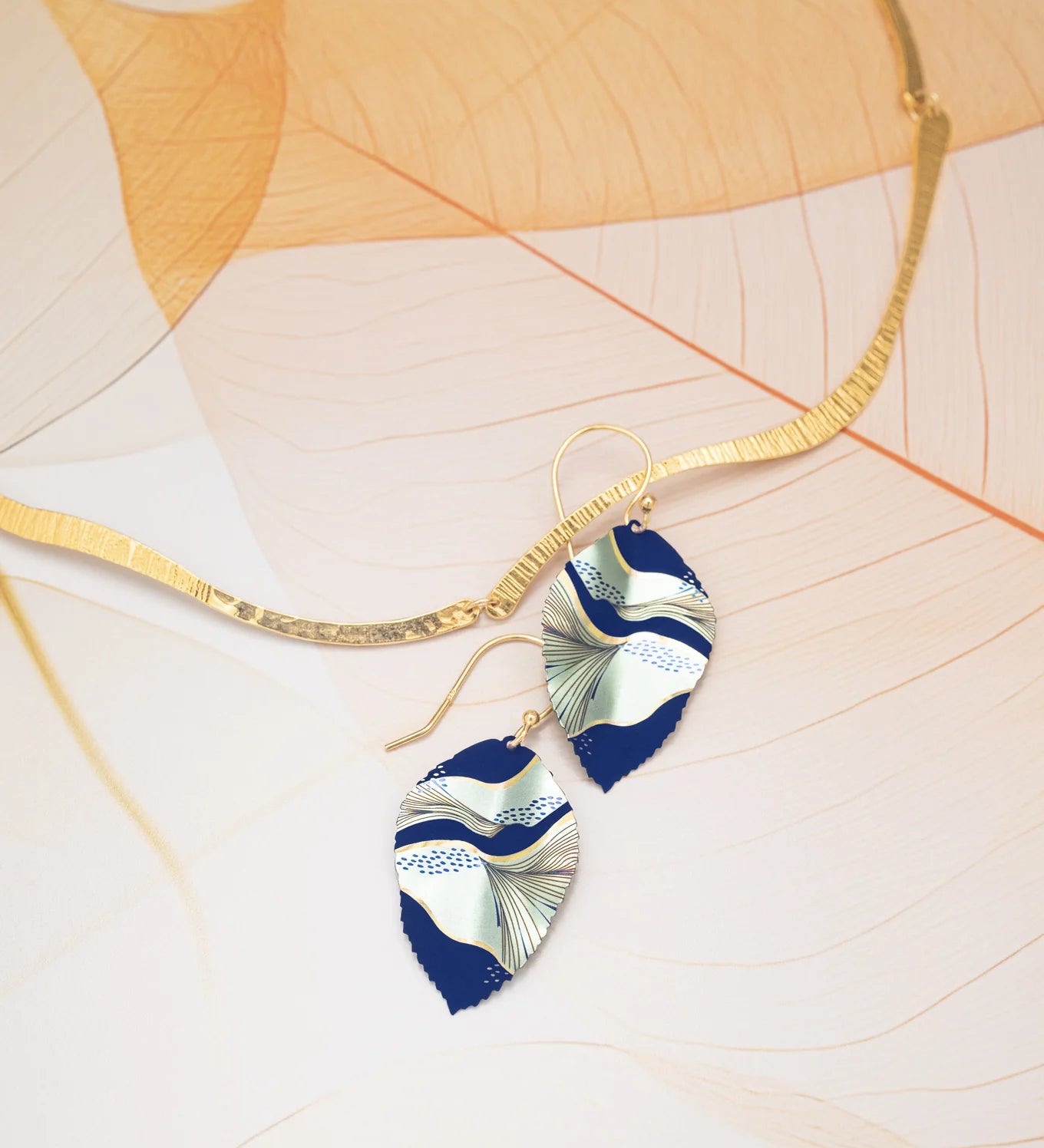 A gold, wavy-textured necklace sits beside Holly Yashi's Eccentric Elm Indigo Earrings—handcrafted leaf earrings in navy blue and white niobium with abstract patterns and gold-filled wires—on a delicate, translucent leaf background.