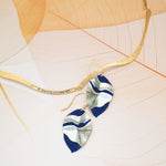 A gold, wavy-textured necklace sits beside Holly Yashi's Eccentric Elm Indigo Earrings—handcrafted leaf earrings in navy blue and white niobium with abstract patterns and gold-filled wires—on a delicate, translucent leaf background.