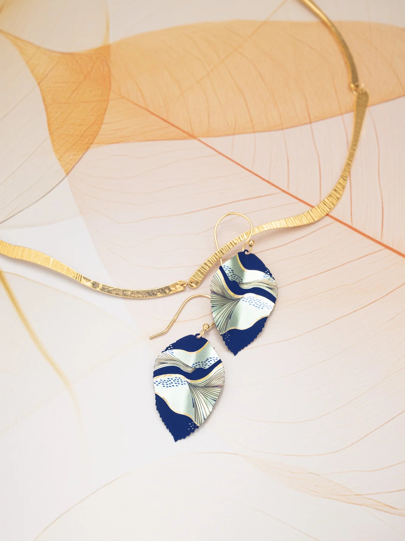 A gold, wavy-textured necklace sits beside Holly Yashi's Eccentric Elm Indigo Earrings—handcrafted leaf earrings in navy blue and white niobium with abstract patterns and gold-filled wires—on a delicate, translucent leaf background.