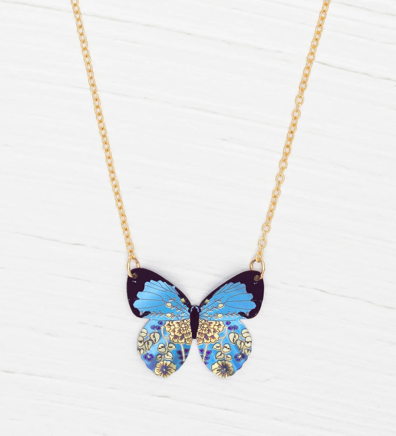The Holly Yashi Blooming Butterfly Blue Radiance Necklace features an iridescent niobium butterfly pendant with gold and floral accents, shown on a white wooden surface.