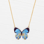 The Holly Yashi Blooming Butterfly Blue Radiance Necklace features an iridescent niobium butterfly pendant with gold and floral accents, shown on a white wooden surface.