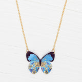The Holly Yashi Blooming Butterfly Blue Radiance Necklace features an iridescent niobium butterfly pendant with gold and floral accents, shown on a white wooden surface.
