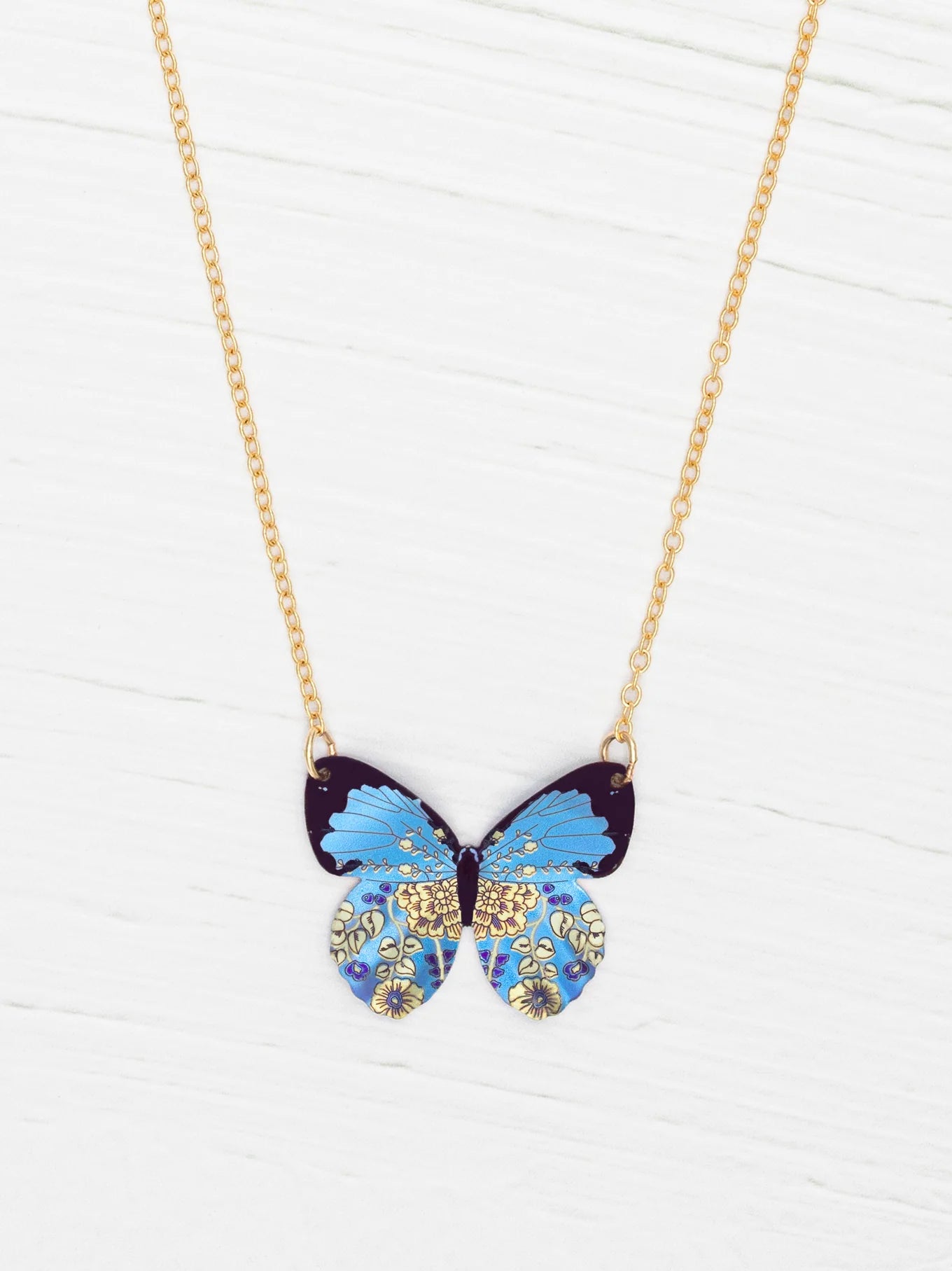 The Holly Yashi Blooming Butterfly Blue Radiance Necklace features an iridescent niobium butterfly pendant with gold and floral accents, shown on a white wooden surface.