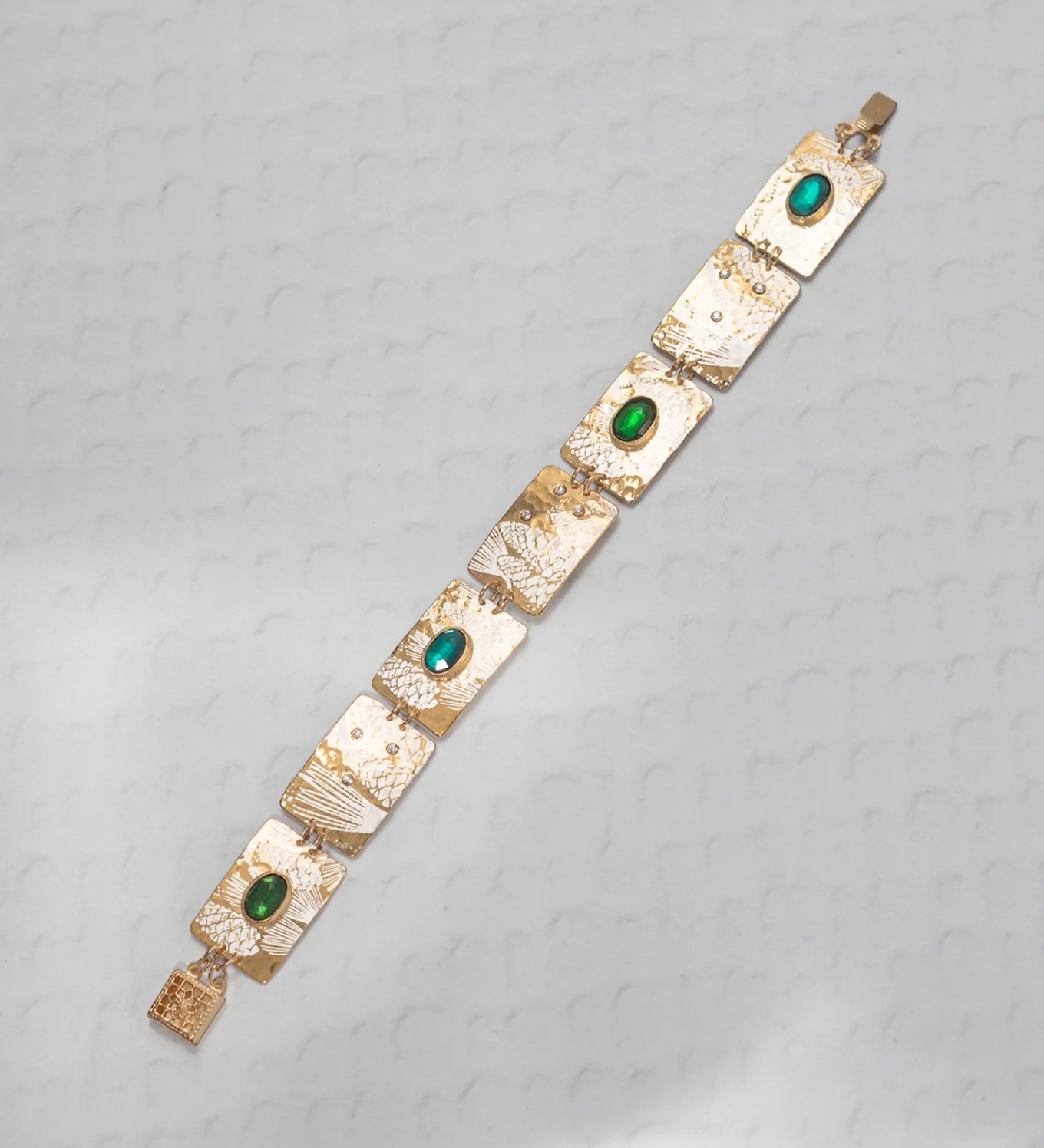 The Gilded Petra Emerald Bracelet by Holly Yashi showcases rectangular textured links adorned with oval emerald green crystals. The handcrafted bracelet is shown diagonally on a light gray patterned surface.