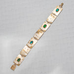 The Gilded Petra Emerald Bracelet by Holly Yashi showcases rectangular textured links adorned with oval emerald green crystals. The handcrafted bracelet is shown diagonally on a light gray patterned surface.