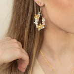A woman with long, light brown hair wears Holly Yashi's Rafaela Gold & White Earrings. Her hand rests near her cheek, partially obscuring her face, while gold and white accents catch the light.