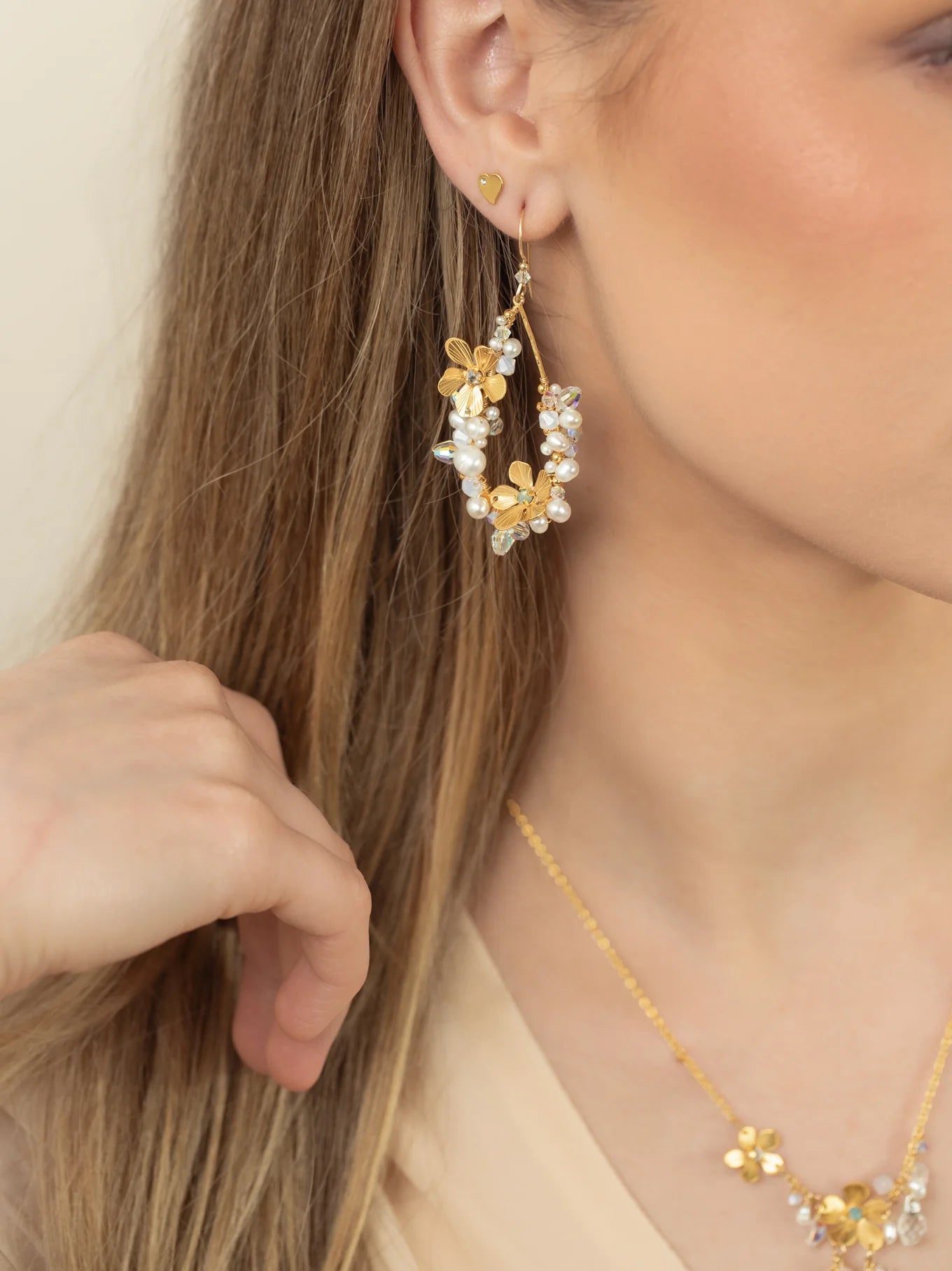 A woman with long, light brown hair wears Holly Yashi's Rafaela Gold & White Earrings. Her hand rests near her cheek, partially obscuring her face, while gold and white accents catch the light.