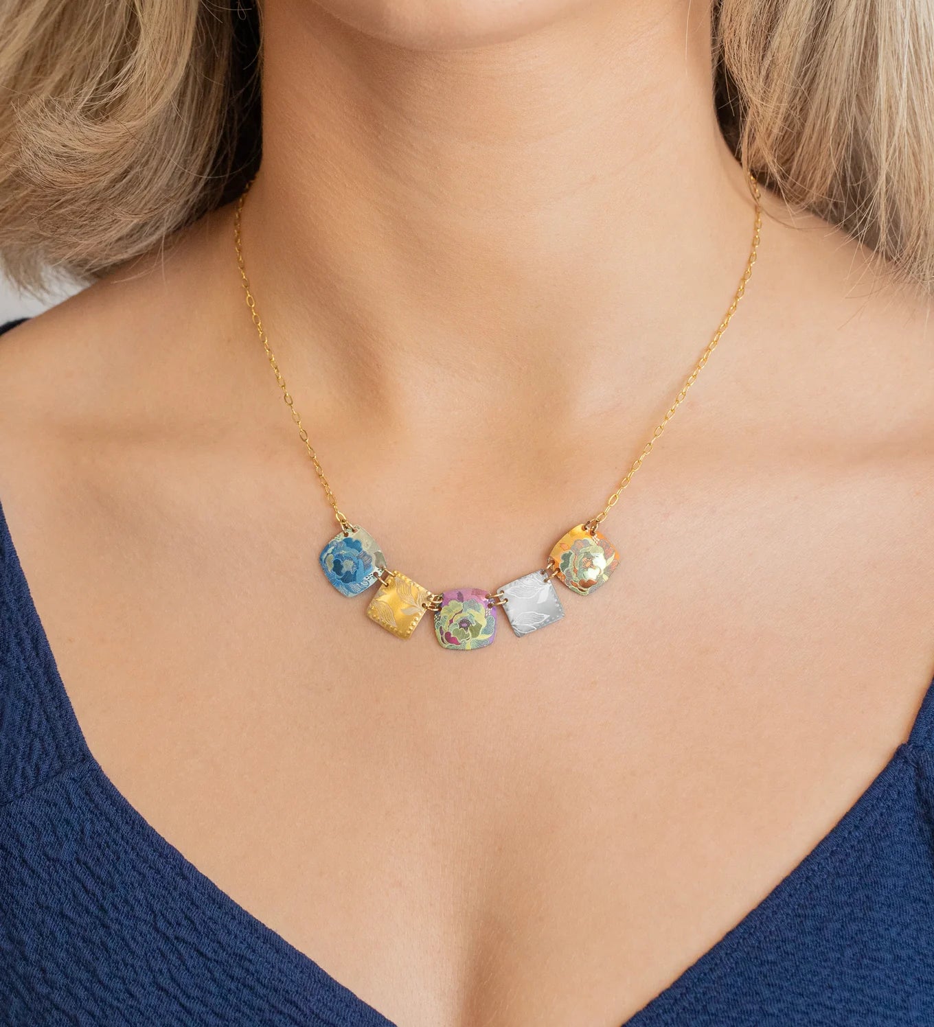A woman wearing the Holly Yashi Flora Multi Color Necklace—featuring five colorful, cube-shaped glass pendants on a gold chain—with a deep blue V-neck top.