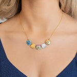 A woman wearing the Holly Yashi Flora Multi Color Necklace—featuring five colorful, cube-shaped glass pendants on a gold chain—with a deep blue V-neck top.