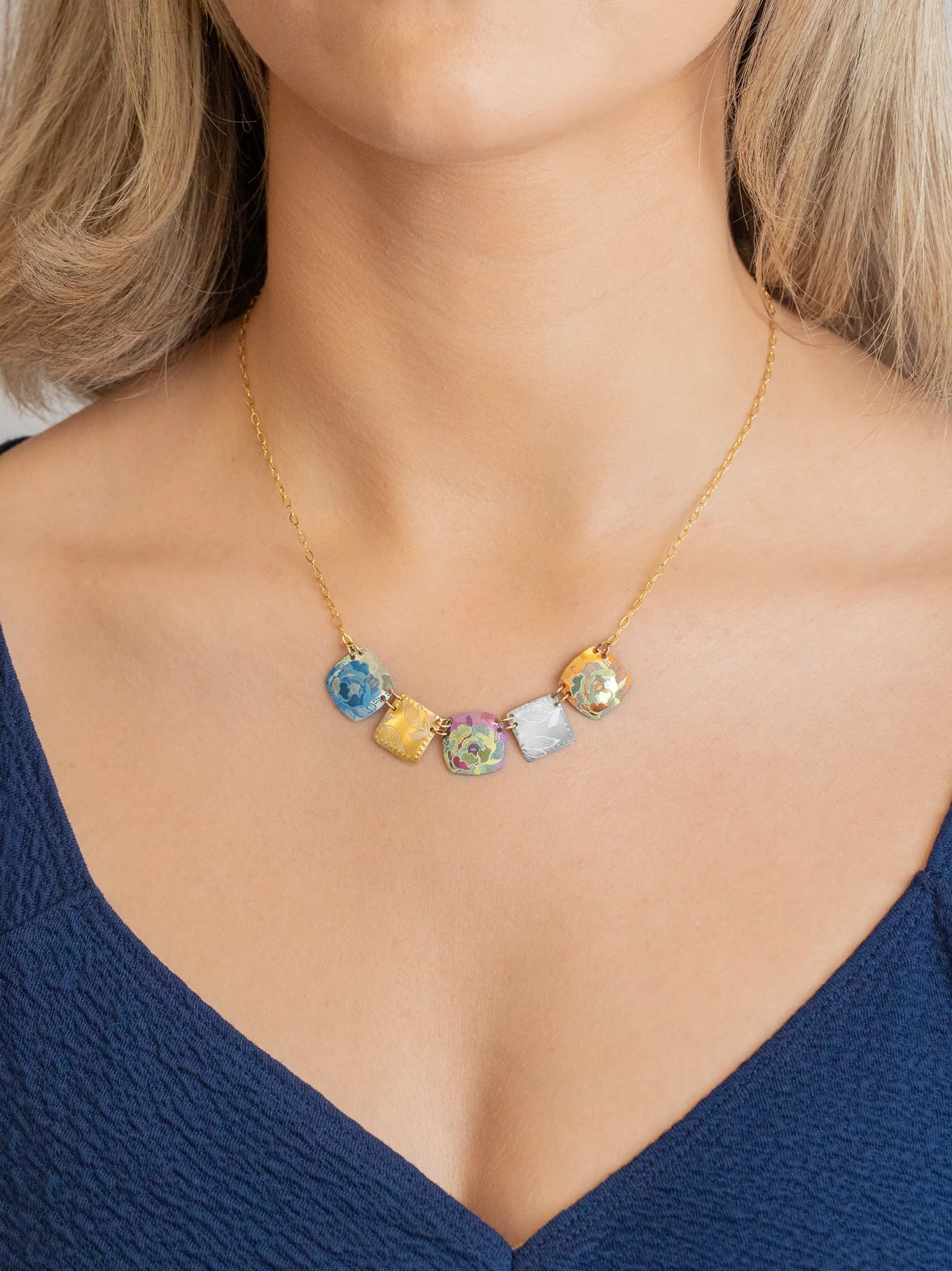 A woman wearing the Holly Yashi Flora Multi Color Necklace—featuring five colorful, cube-shaped glass pendants on a gold chain—with a deep blue V-neck top.