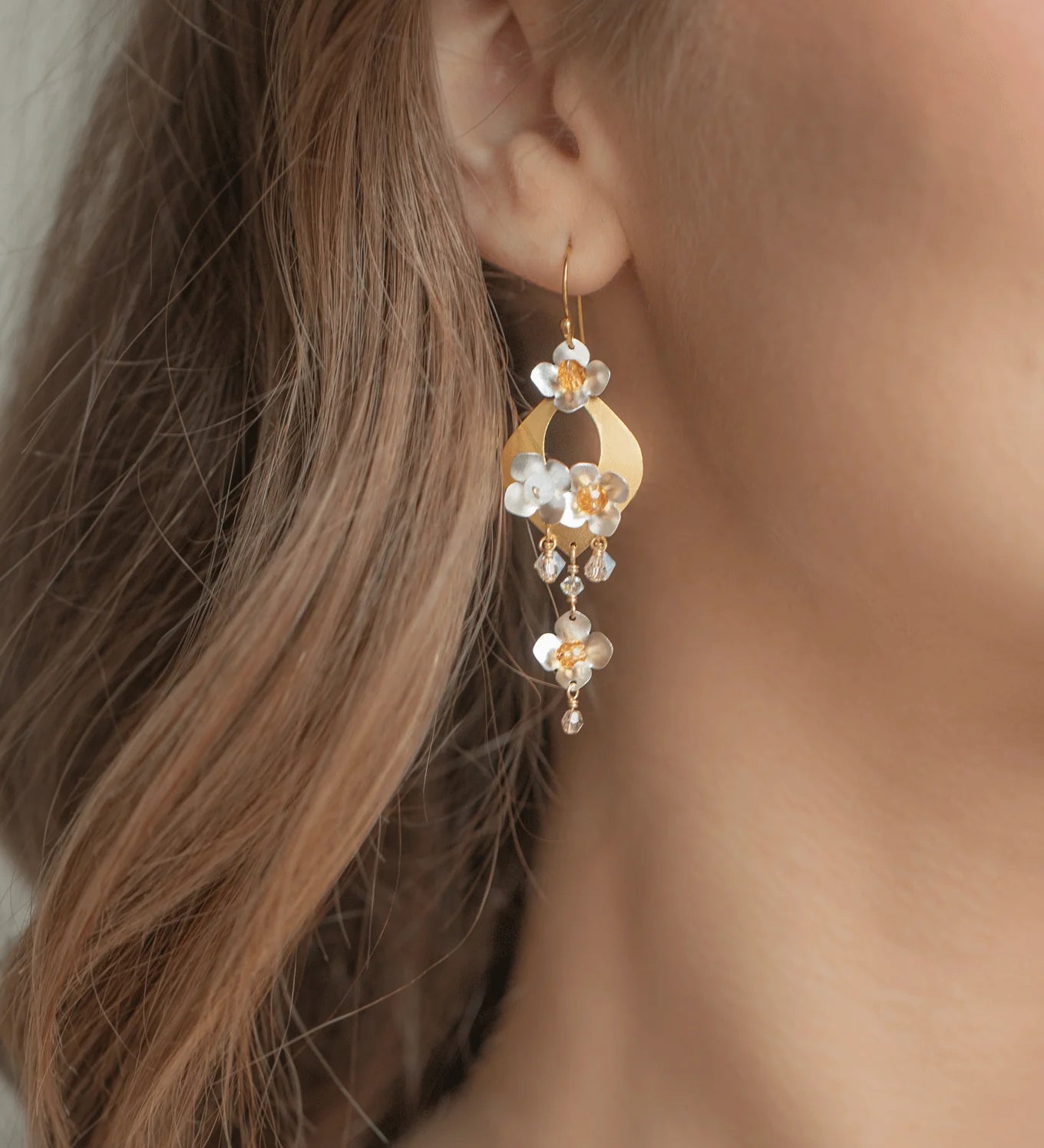 Close-up of a woman's ear wearing Holly Yashi's Hawaiian Wedding Gold/Silver Earrings—dangle earrings with white and amber flower-shaped beads. Her wavy brown hair beautifully frames the elegant Holly Yashi jewelry.