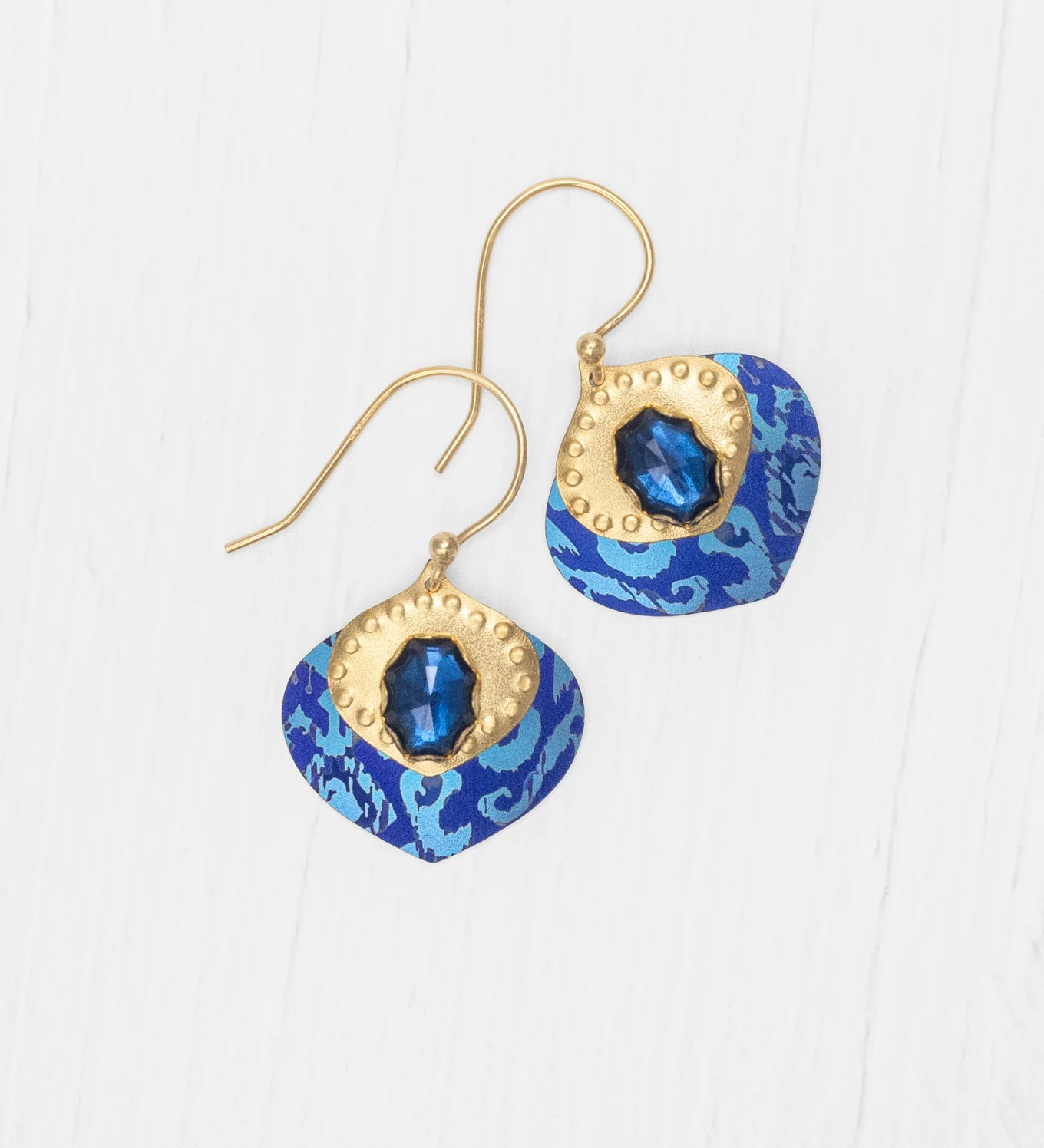 The Arabella Sapphire Blue Earrings by Holly Yashi feature gold hooks, blue and gold accents, a central shiny blue gem, and a patterned blue background displayed on a white wood surface.