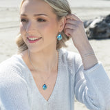A blonde woman smiles on a sandy beach, wearing a light sweater, silver bracelets, and the Holly Yashi Lani/Kai Blue Necklace with matching earrings. Her hypoallergenic jewelry sparkles as the ocean and rocks shimmer behind her.