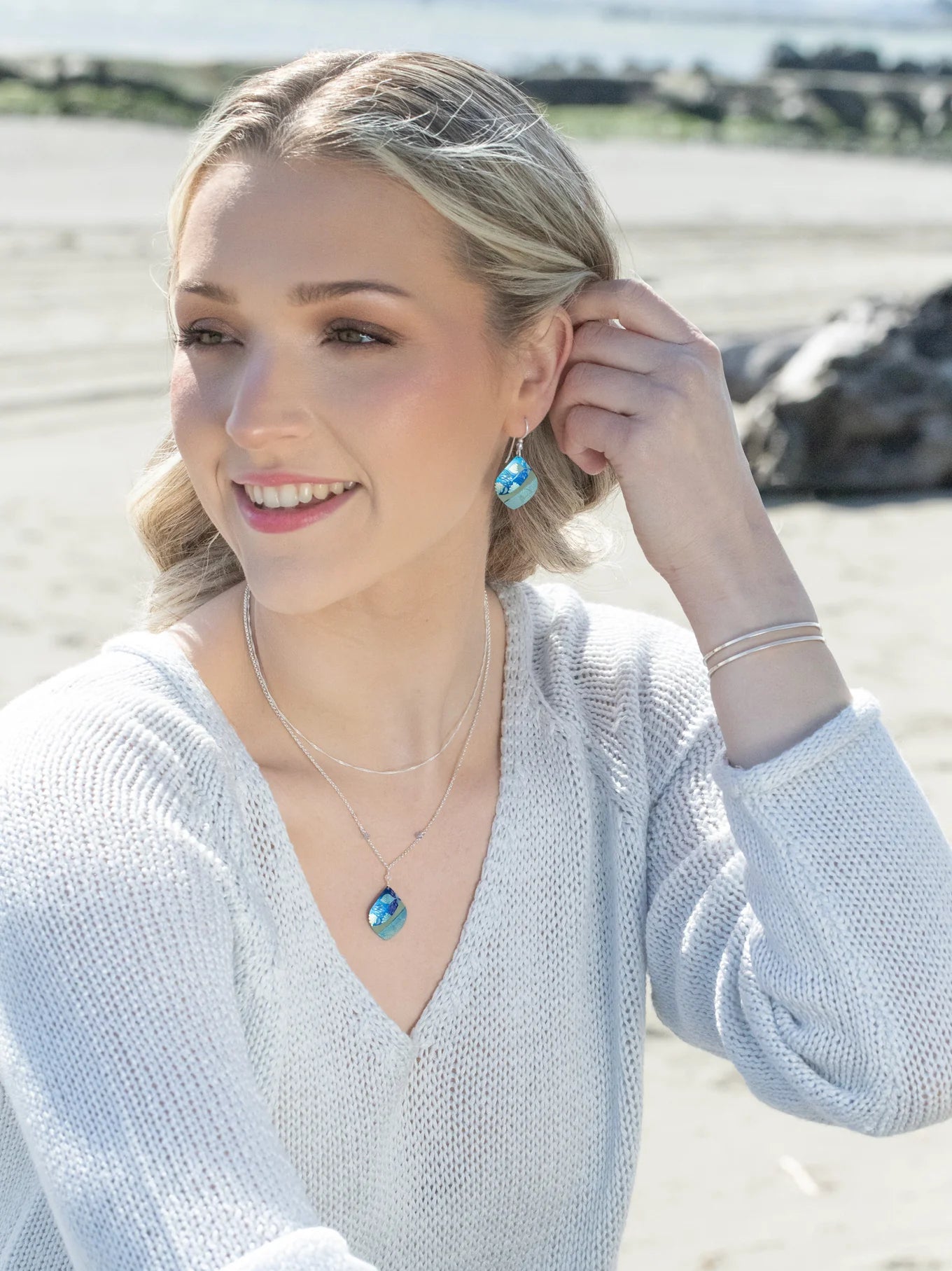 A blonde woman smiles on a sandy beach, wearing a light sweater, silver bracelets, and the Holly Yashi Lani/Kai Blue Necklace with matching earrings. Her hypoallergenic jewelry sparkles as the ocean and rocks shimmer behind her.