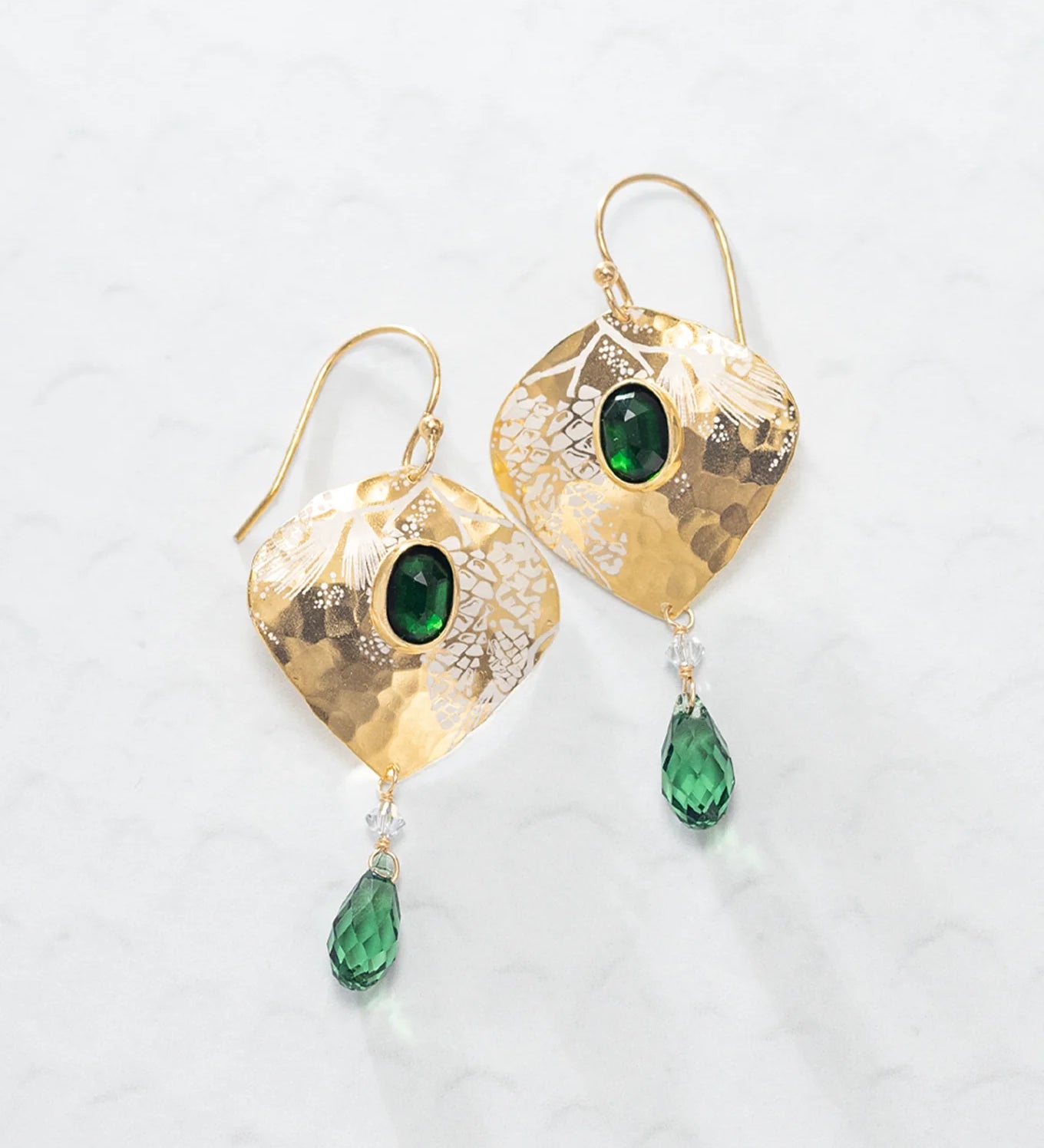 The Estelle Emerald Earrings by Holly Yashi feature hammered gold ovals with a central green gemstone and a dangling green teardrop, shown on a light textured surface.