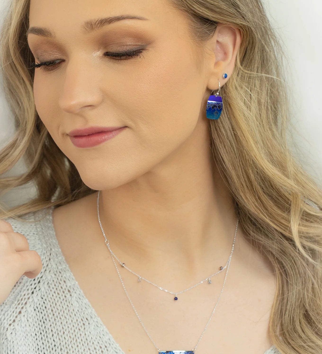 A woman with long blonde hair wears a light knit top and handcrafted Holly Yashi jewelry—a Magnolia Midnight Blue Necklace and blue dangle earrings. She looks down slightly with a soft smile and natural makeup.