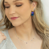 A woman with long blonde hair wears a light knit top and handcrafted Holly Yashi jewelry—a Magnolia Midnight Blue Necklace and blue dangle earrings. She looks down slightly with a soft smile and natural makeup.