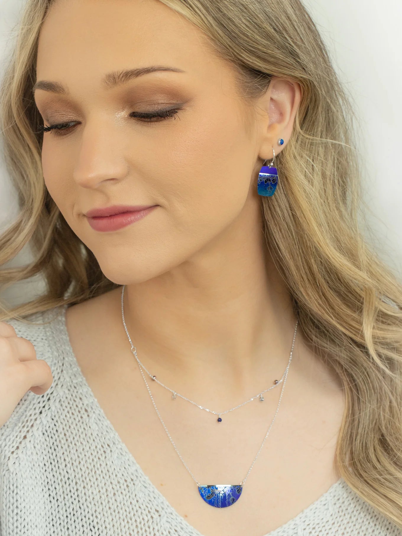 A woman with long blonde hair wears a light knit top and handcrafted Holly Yashi jewelry—a Magnolia Midnight Blue Necklace and blue dangle earrings. She looks down slightly with a soft smile and natural makeup.