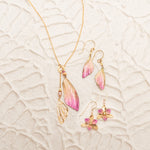 The Holly Yashi Special Edition Flutterby Pink Lariat Necklace and two pairs of matching earrings—one with pink and gold wing motifs, the other with pink flowers—are displayed on a textured, light surface.