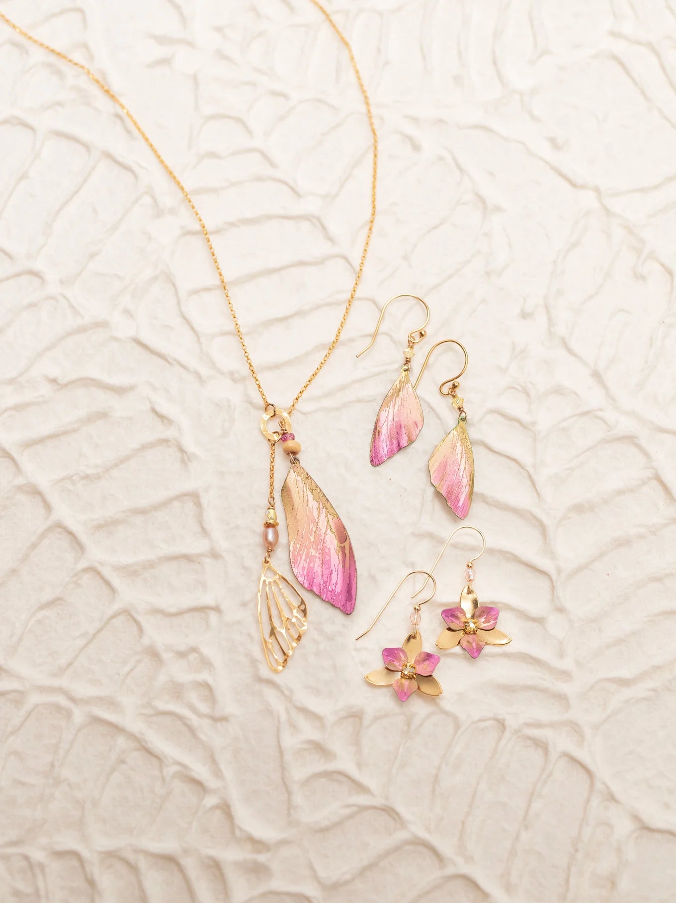 The Holly Yashi Special Edition Flutterby Pink Lariat Necklace and two pairs of matching earrings—one with pink and gold wing motifs, the other with pink flowers—are displayed on a textured, light surface.
