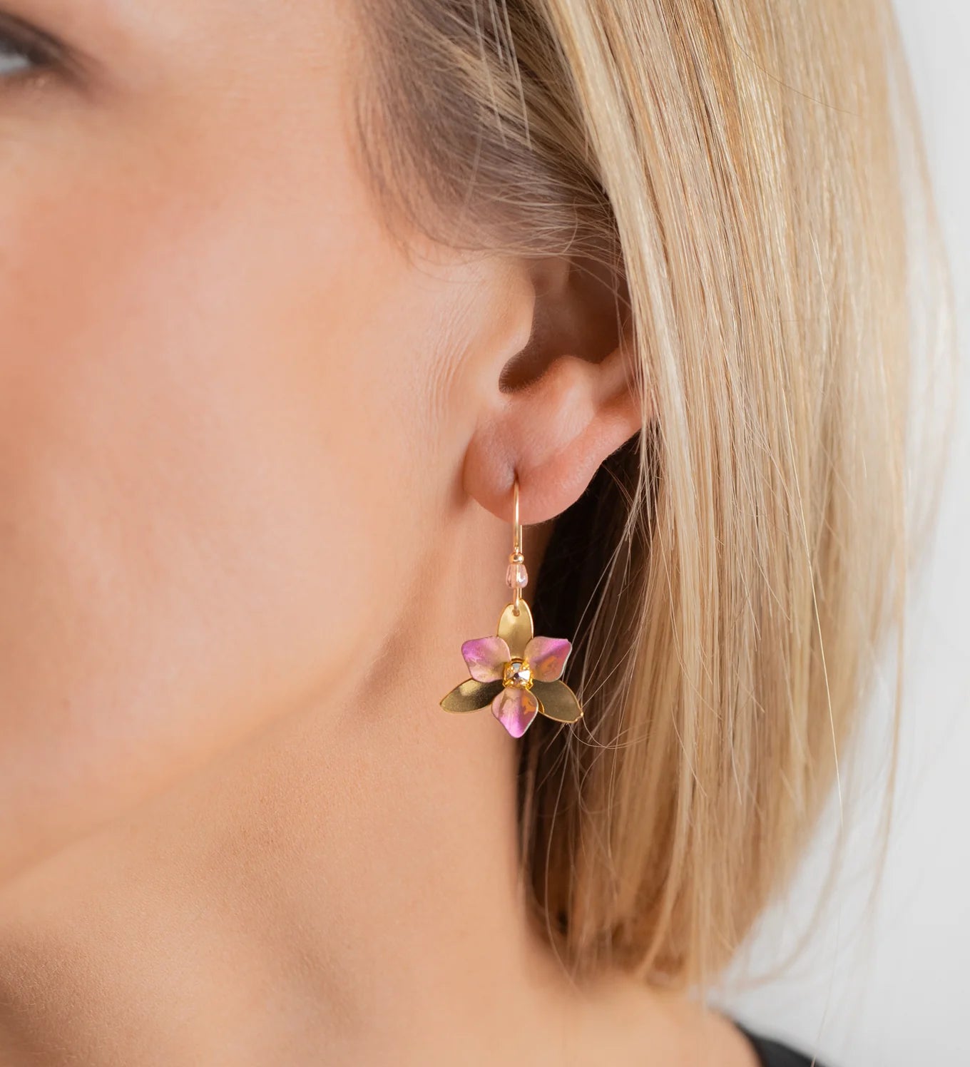 A close-up of a womans ear wearing the Special Edition Orla Drops Earrings by Holly Yashi, featuring dangling flower-shaped designs with pink Austrian crystal petals and green leaves. She has straight blonde hair and light skin.