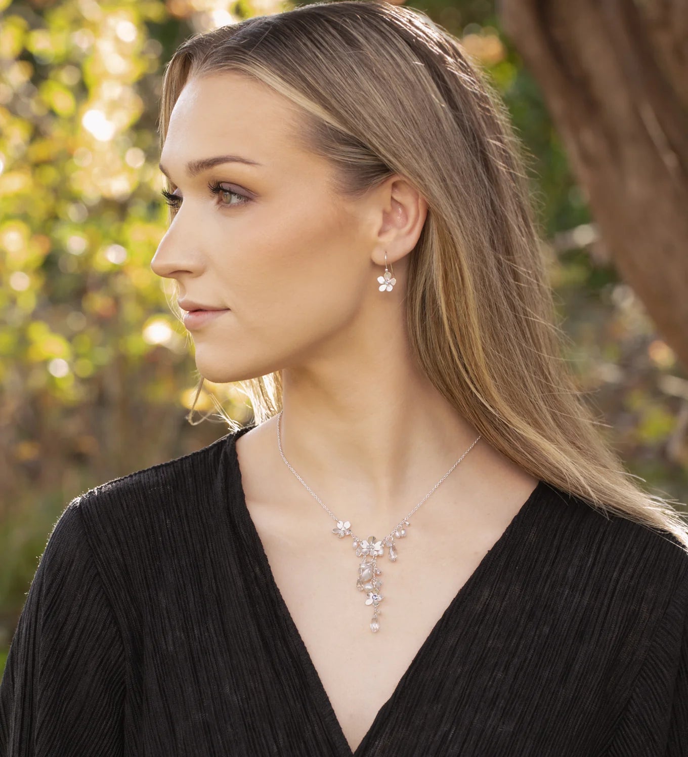 A woman with long blonde hair in a black textured top gazes sideways outdoors, wearing the Rafaela Gold & White Necklace by Holly Yashi and matching earrings. Sunlit trees and greenery create a softly blurred background.