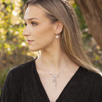 A woman with long blonde hair in a black textured top gazes sideways outdoors, wearing the Rafaela Gold & White Necklace by Holly Yashi and matching earrings. Sunlit trees and greenery create a softly blurred background.