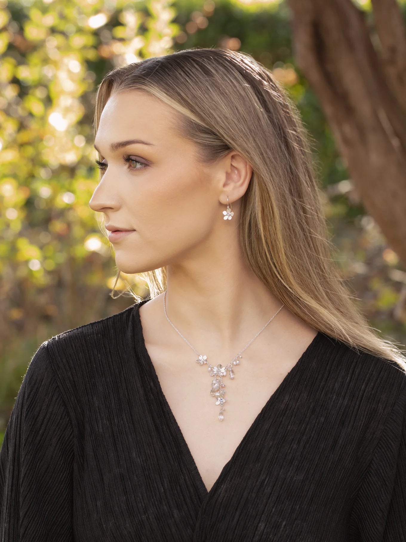 A woman with long blonde hair in a black textured top gazes sideways outdoors, wearing the Rafaela Gold & White Necklace by Holly Yashi and matching earrings. Sunlit trees and greenery create a softly blurred background.
