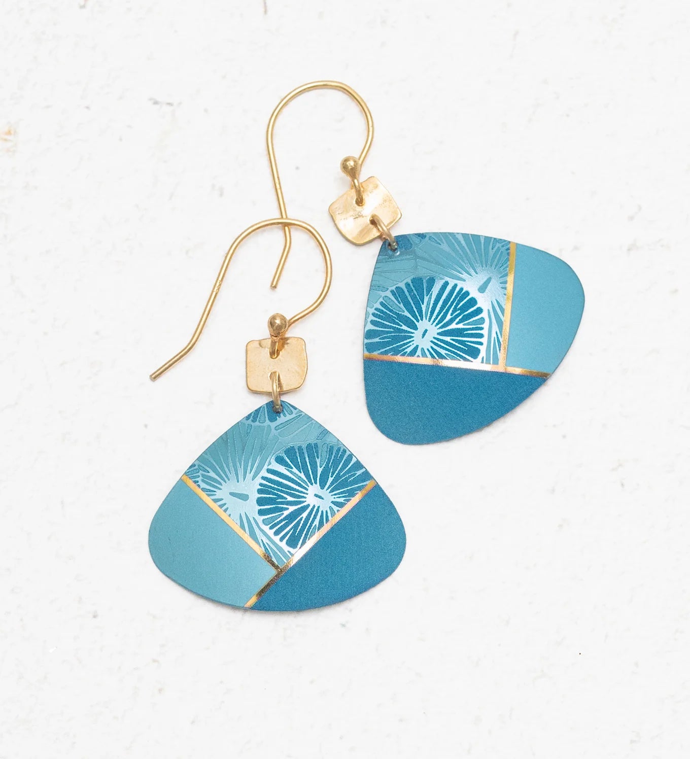 The Rowan Sky Blue Earrings by Holly Yashi feature gold hooks, geometric blue pendants, and hypoallergenic materials. Their abstract light blue, teal, and gold accents are highlighted with delicate white floral designs.