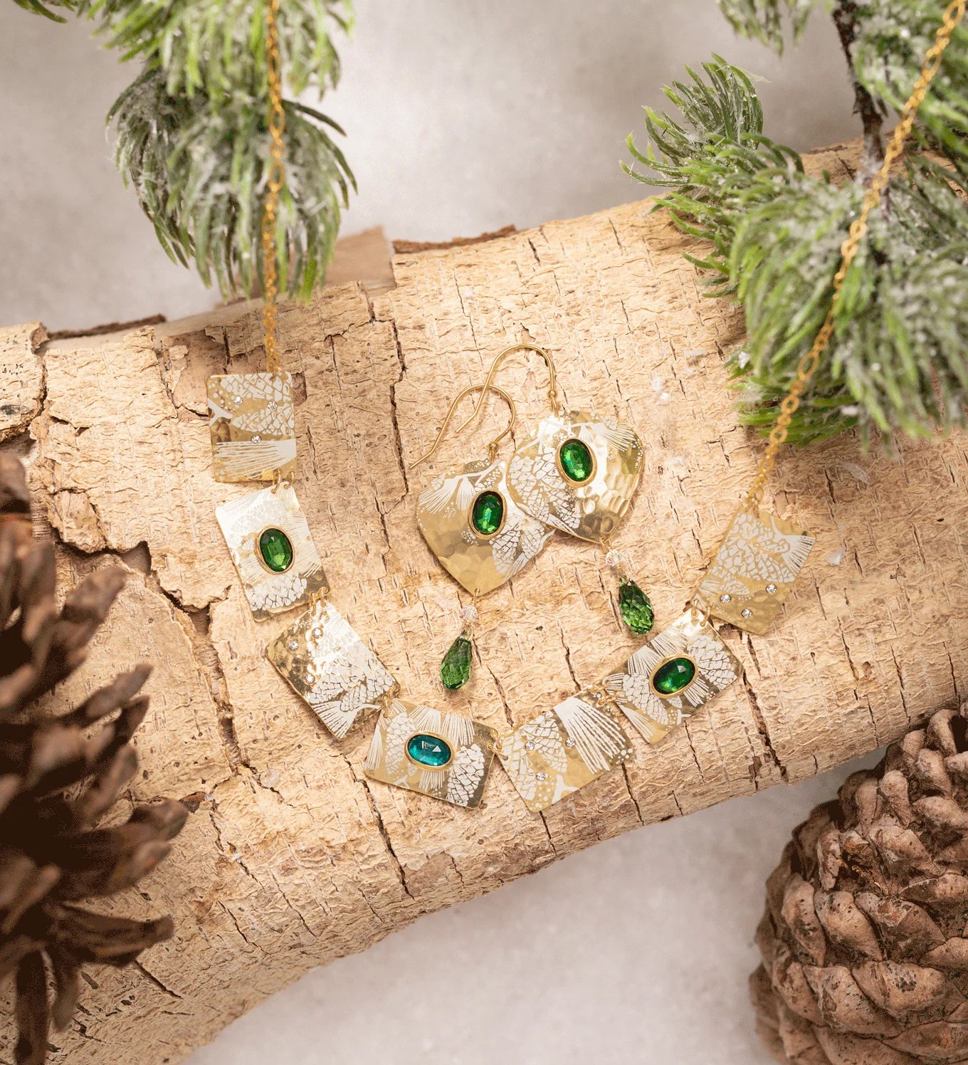 The Estelle Emerald Earrings by Holly Yashi and matching necklace are showcased on a light wood log with pine branches and pinecones, set against a snowy backdrop.