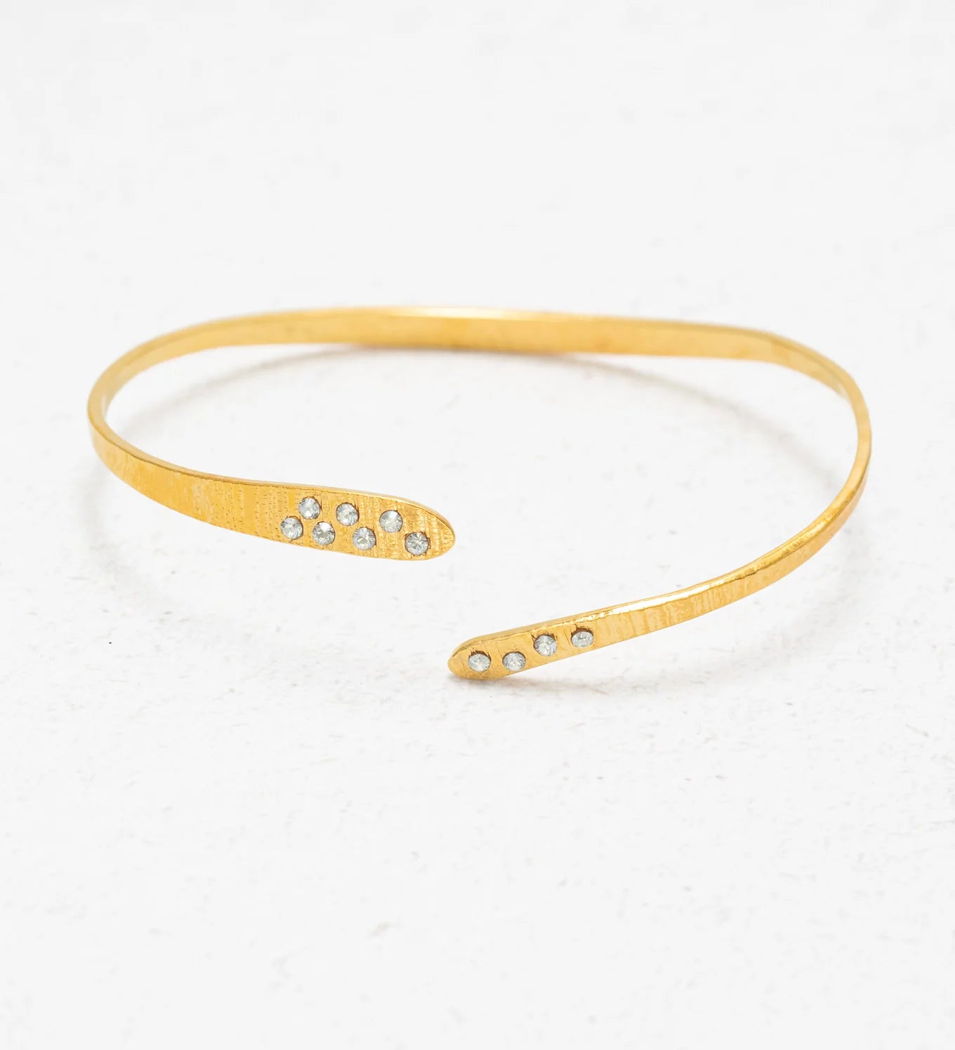 The Holly Yashi Zara Cuff Gold Bracelet is a thin, open-ended gold piece featuring small clear gemstones at each tip, shown on a white textured surface.