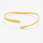 The Holly Yashi Zara Cuff Gold Bracelet is a thin, open-ended gold piece featuring small clear gemstones at each tip, shown on a white textured surface.