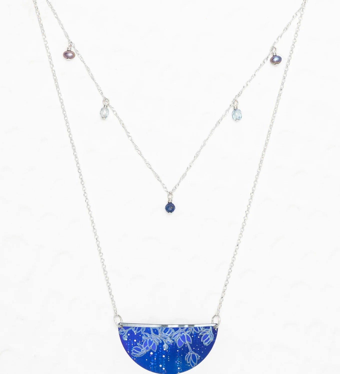 The Magnolia Midnight Blue Necklace by Holly Yashi features a semicircular blue gemstone pendant with a white floral pattern and a second chain with small, colored gemstones—an elegant piece of handcrafted jewelry.