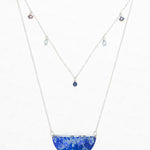 The Magnolia Midnight Blue Necklace by Holly Yashi features a semicircular blue gemstone pendant with a white floral pattern and a second chain with small, colored gemstones—an elegant piece of handcrafted jewelry.