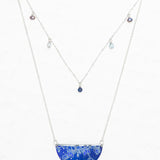 The Magnolia Midnight Blue Necklace by Holly Yashi features a semicircular blue gemstone pendant with a white floral pattern and a second chain with small, colored gemstones—an elegant piece of handcrafted jewelry.