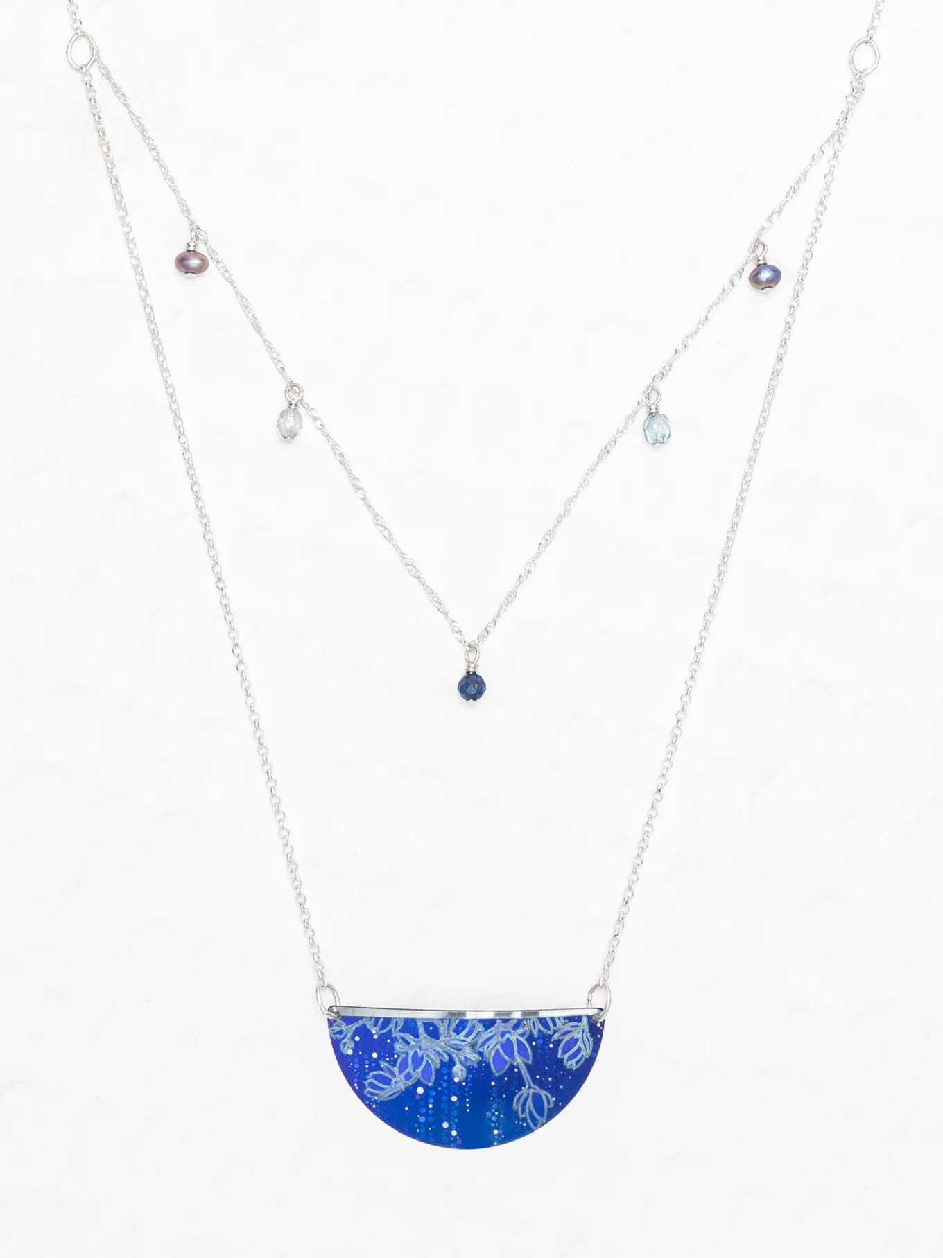 The Magnolia Midnight Blue Necklace by Holly Yashi features a semicircular blue gemstone pendant with a white floral pattern and a second chain with small, colored gemstones—an elegant piece of handcrafted jewelry.