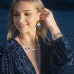 A woman in a Midnight Blue textured dress smiles with closed eyes, touching her Holly Yashi Aster Earrings in Midnight Blue. She wears a matching necklace and hypoallergenic bracelet while standing outdoors near the water at dusk.
