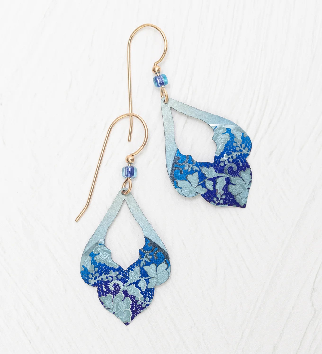 The Marisol Forever Blue Earrings by Holly Yashi feature gold hooks, hypoallergenic niobium wires, blue beads, and layered teardrop pendants with a blue floral pattern on light blue, shown on a white textured surface.