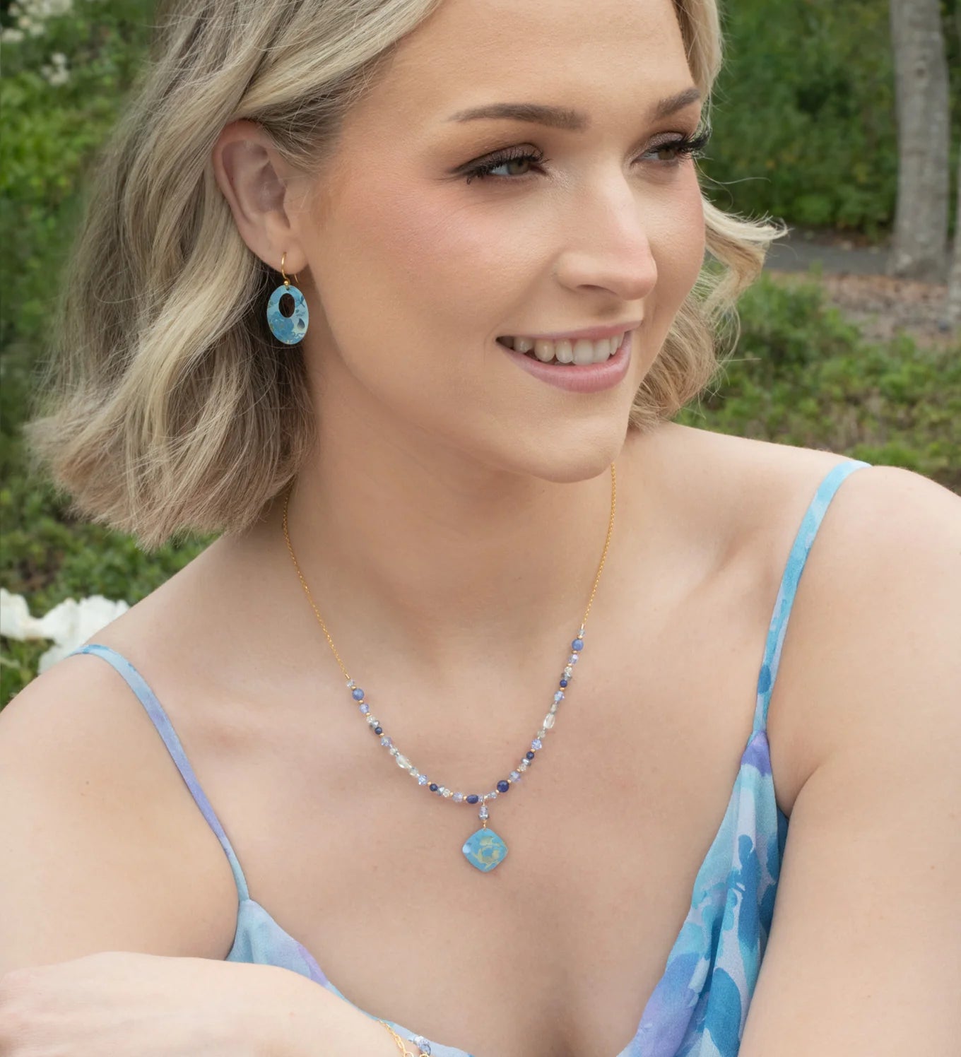 A smiling woman with short blonde hair, in a blue and purple dress, sits outdoors among flowers. She wears matching blue earrings and a striking Elena Beaded Necklace Sky Blue Necklace by Holly Yashi.