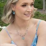 A smiling woman with short blonde hair, in a blue and purple dress, sits outdoors among flowers. She wears matching blue earrings and a striking Elena Beaded Necklace Sky Blue Necklace by Holly Yashi.