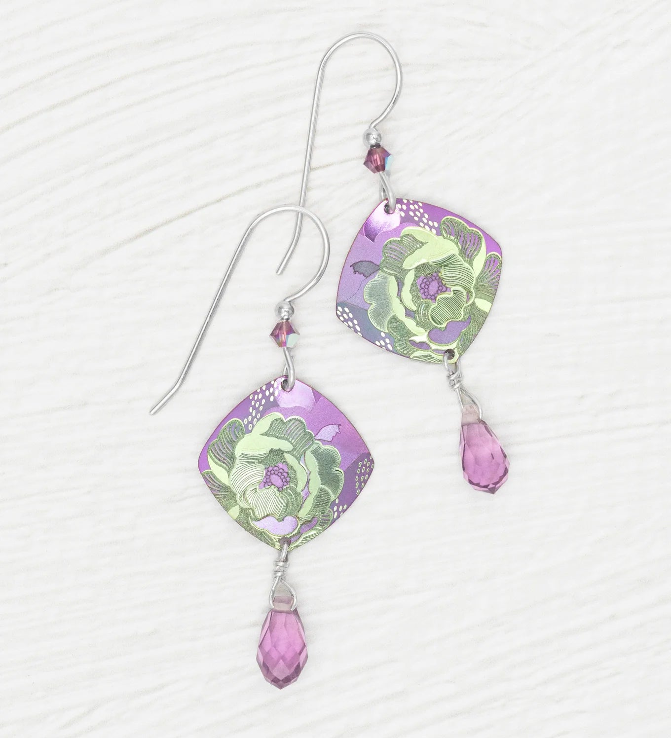 A pair of Elena Violet Earrings by Holly Yashi, featuring purple square-shaped charms with green floral designs and small purple crystal drops, displayed on a white textured surface.