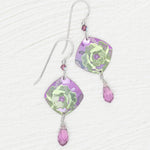 A pair of Elena Violet Earrings by Holly Yashi, featuring purple square-shaped charms with green floral designs and small purple crystal drops, displayed on a white textured surface.