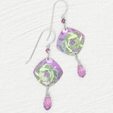 A pair of Elena Violet Earrings by Holly Yashi, featuring purple square-shaped charms with green floral designs and small purple crystal drops, displayed on a white textured surface.