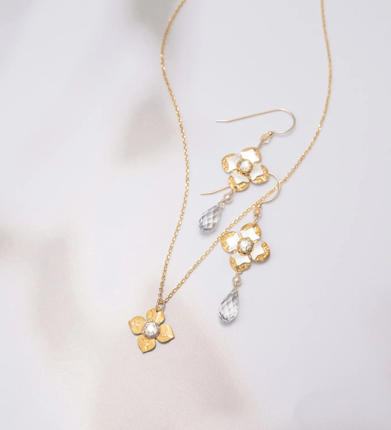 Octavia Gold Silver Earrings by Holly Yashi, inspired by Victorian style, feature clear teardrop crystals and are shown with a gold flower pendant necklace on a soft, light background.