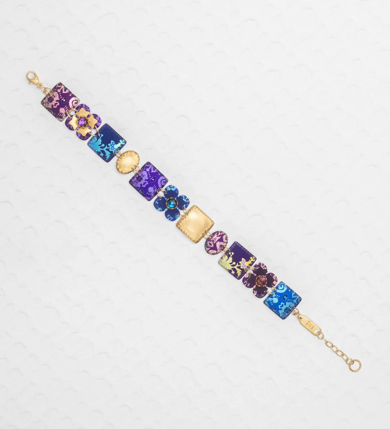 The Eliza Tiled Multi-Color Bracelet by Holly Yashi features a gold chain with square and round charms in purple, blue, and pink, each adorned with floral and abstract designs, displayed on a white textured surface.