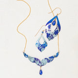 The Isla Forever Blue Necklace by Holly Yashi is a hypoallergenic jewelry set with a gold chain, blue and silver floral pendants, and teardrop blue gemstones, shown on a light textured background.