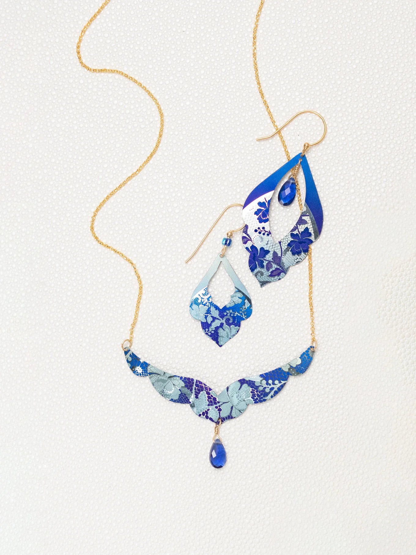 The Isla Forever Blue Necklace by Holly Yashi is a hypoallergenic jewelry set with a gold chain, blue and silver floral pendants, and teardrop blue gemstones, shown on a light textured background.